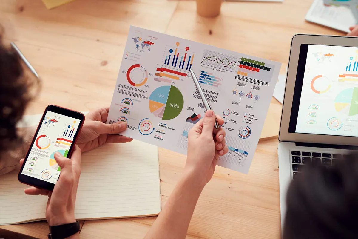 Manos revisando gráficas de resultados en papel, en el celular y en el portátil